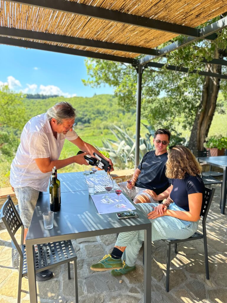 Winemaker pouring red wine for a couple on a terrace overlooking Chianti Classico vineyards in Tuscany.