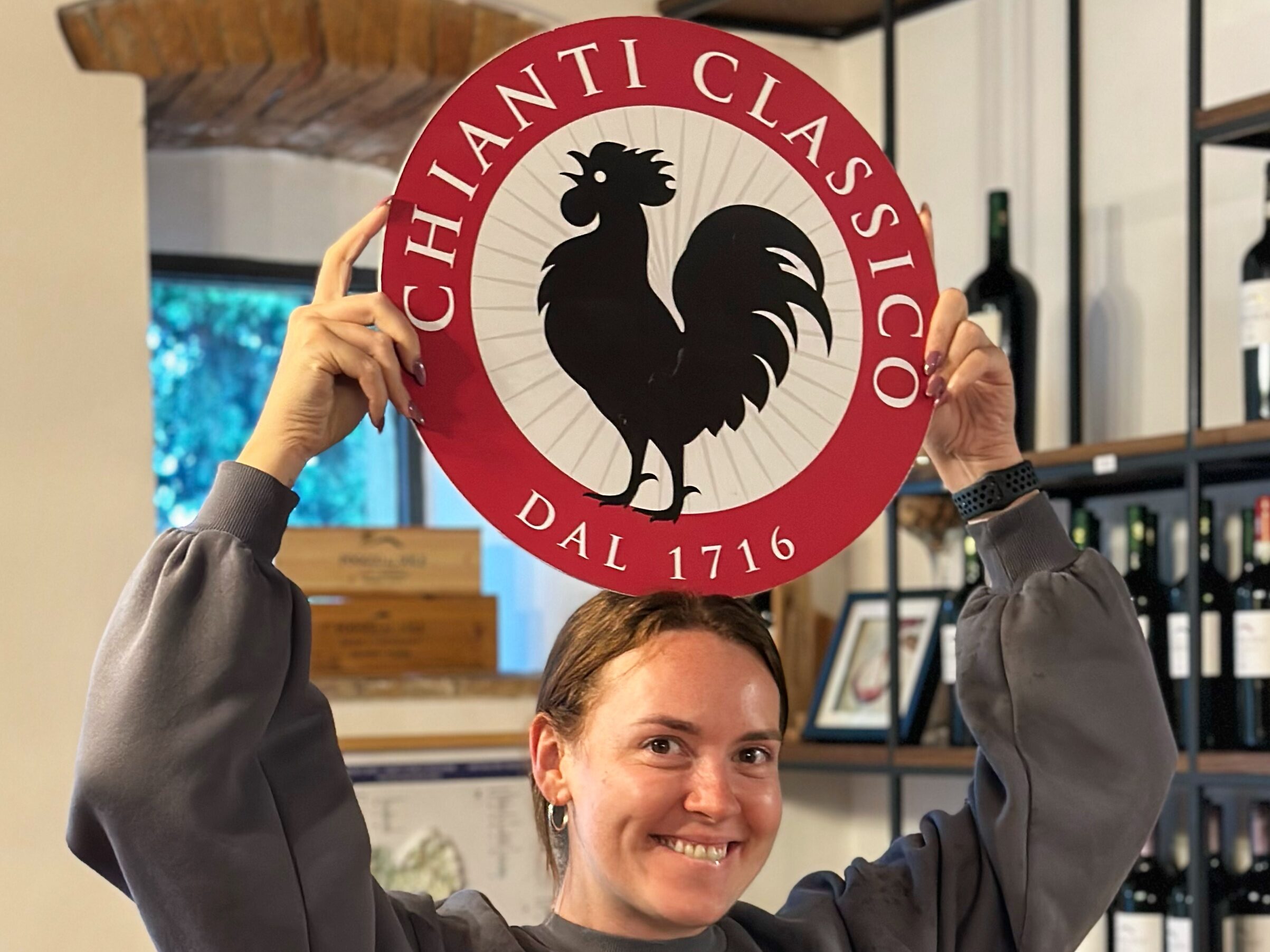 Tuscany Uncorked guest holding the Chianti Classico Black Rooster sign in a winery tasting room during a wine tour from Florence.