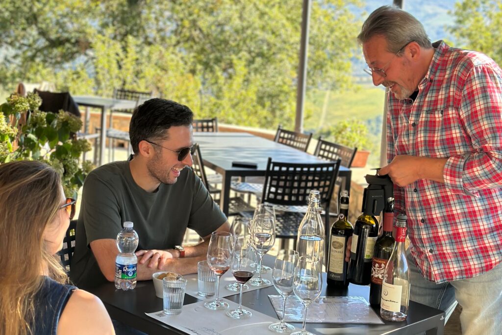 Two guests smiling during a wine tasting on a Tuscany wine tour from Florence, seated with vineyard views in the background.