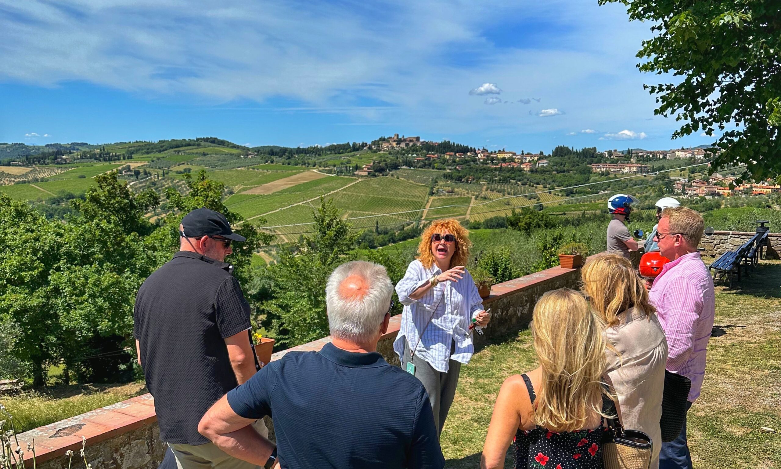 Small group of travelers listening to a guide during a scenic stop on a Tuscany wine tour, with vineyard-covered hills and a hilltop village in the background.