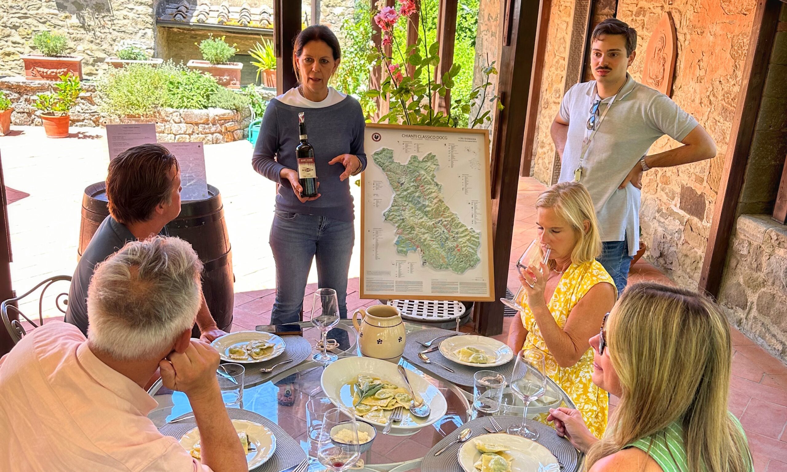 How Many Wineries Can You Visit in One Day in Tuscany? The answer is in this picture where you can see guests enjoying a traditional Tuscan lunch and guided wine tasting at a family-run winery, with a producer presenting Chianti Classico wines and the wine region map.